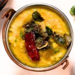 Top view of a bowl filled with cooked yellow lentils, topped with curry leaves, cumin seeds, and a red chile.